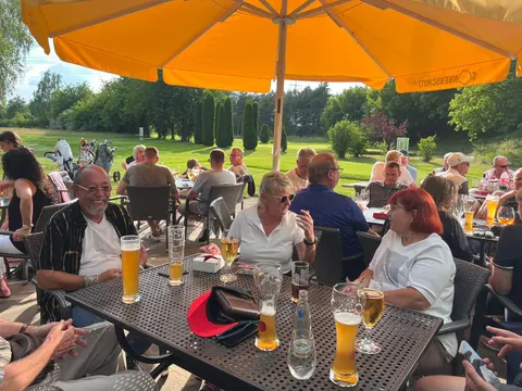 Gäste sitzen unter einem Sonnenschirm auf der Clubhausterrasse mit Blick auf den Platz