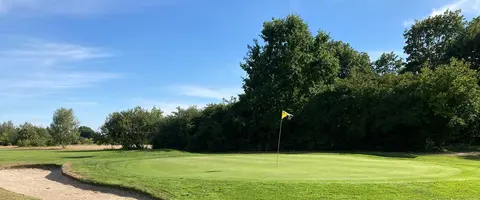 Grün mit Fahne und Bunker unter blauem Himmel, umgeben von Laubbäumen